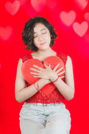 Valentine's day, young girl holds a red paper heart, she makes cheerful expressions, concept of love.の写真素材