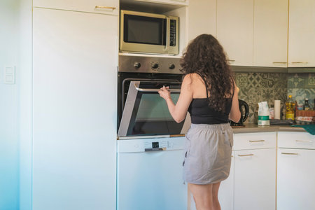 Middle-aged woman in her kitchen baking, she is opening the oven, in her kitchen the woman opens the oven to take out her food.の写真素材