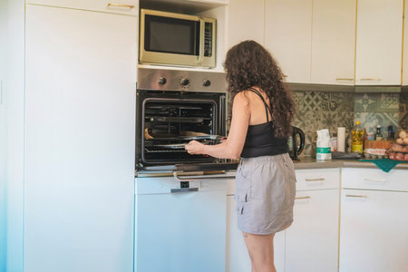 Middle-aged woman in her kitchen baking, she is opening the oven, in her kitchen the woman opens the oven to take out her food.の写真素材