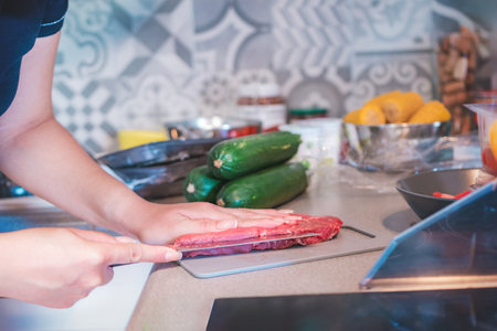 Photograph of a woman cooking in her kitchen. She is cutting meat with a sharp knife. Her kitchen is full of items for preparing meat, including vegetables ready to be chopped.の写真素材