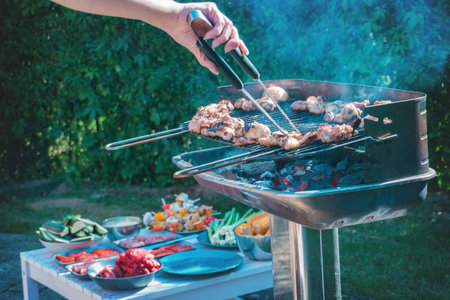 A grill full of meat roasting, the grill lit, and only a pair of hands with tongs are shown, turning...