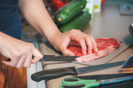 Photograph of a woman cooking in her kitchen. She is cutting meat with a sharp knife. Her kitchen is full of items for preparing meat, including vegetables ready to be chopped.の写真素材