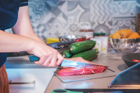 Photograph of a woman cooking in her kitchen. She is cutting meat with a sharp knife. Her kitchen is full of items for preparing meat, including vegetables ready to be chopped.の写真素材