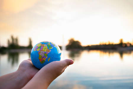 Detail of person's hands holding small ball of the world in front of a lake at sunset. Selective focus. Concept of travel and care of the planet.の写真素材