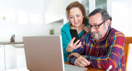 Elderly couple using simultaneously their smartphone and laptop from their kitchen at home. Stay...