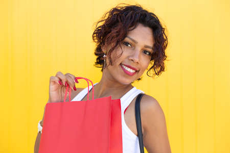 Young woman with brown skin smiling as she shows a shopping bag on a yellow wall. Space for text. Shopping, gifts and sales concept.の写真素材