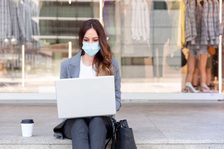 Young businesswoman wearing a face mask outside during the Covid-19 health crisis. She's working with her laptop on the street. Selective focus. Space for text. Business and new normal concept.の写真素材