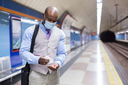 Black man with face mask putting sanitizing gel on his hands. He is inside a subway station. Concept...