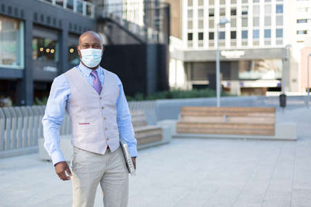 Enterprising black man on the street. He wears a face mask and holds a laptop under his arm. Concept...