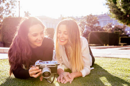 Two women lying on the grass smile as they look at a vintage camera. They have the sun behind them.の写真素材