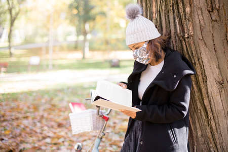 Young woman reading a book in the park. She is wearing winter clothes and a face mask. Space for...