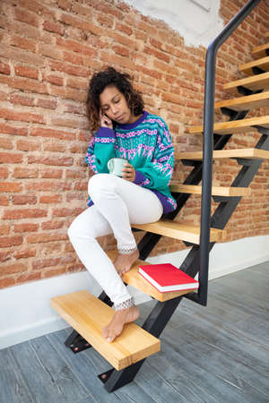 Brunette woman with a thoughtful gesture holding a coffee cup in her hands. She is sitting on the stairs of her house next to a book.の写真素材