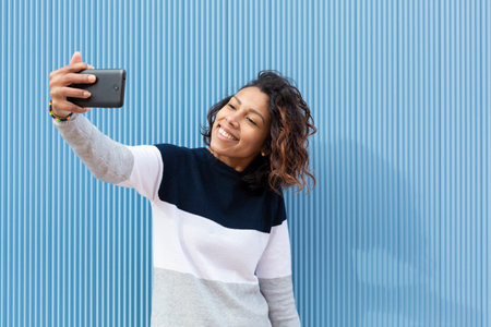 Young and smiling latin woman taking a portrait with her mobile phone on a blue background. Space for text.の写真素材