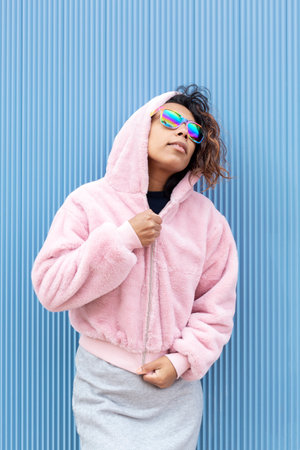 Portrait of a young hispanic woman wearing sunglasses with the colors of the lgbt flag. She is standing isolated on a blue background. Concept of sexual freedom and diversity.の写真素材