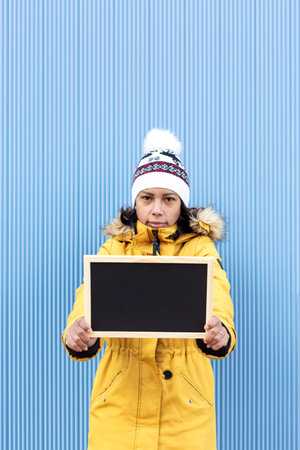 Vertical portrait of a serious latin woman in winter clothing holding a blank chalkboard. She is standing next to a blue wall. Space for text.の写真素材