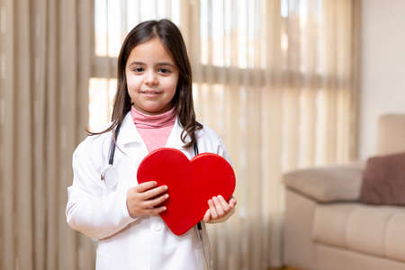 Little child in doctor's uniform holding smiling big heart in her hands. Concept of health and wellness. Space for text.の写真素材
