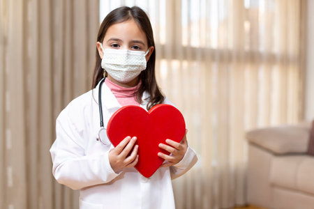 Little girl with doctor's uniform and medical mask holding in her hands a big heart. Concept of health and wellness. Space for text.の写真素材