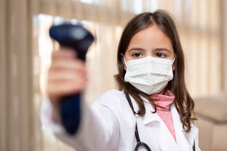 Little girl with doctor's uniform and medical mask using an infrared thermometer. Covid-19 virus safety concept.の写真素材