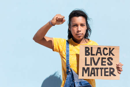 Portrait of Latin American woman with serious attitude and raised fist. She is isolated on blue background holding a banner with slogan Black Lives Matter. Space for text.の写真素材