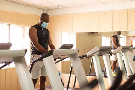 Black man with face mask setting up treadmill in gym. Space for text.