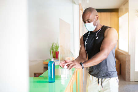 Black man in sportswear and face mask spraying sanitizing gel on his hands. He is entering the gym.