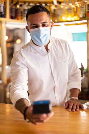 Young latin waiter with a medical mask. He has a dataphone in his hand to make a credit card charge.の写真素材