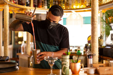 Young Latino bartender preparing a cocktail at the bar. He wears a medical mask.