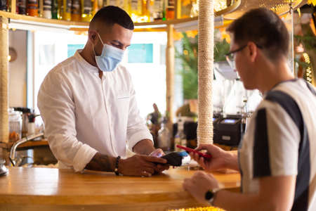 Young Latin American waiter with face mask charging from the bar to a customer by contactless payment.の写真素材