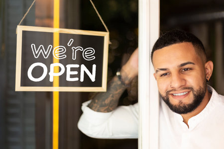 Smiling young Latin American man at the entrance of a small business next to a sign with the text...