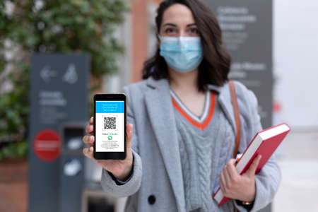 Female student with face mask holding smartphone with digital vaccine passport on the screen....