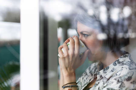 Close up of adult woman drinking coffee. She is at home looking outside through the window.