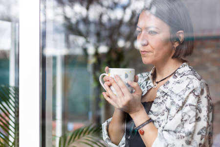 Portrait of adult Caucasian woman with coffee cup in her hands. She is pensively looking through the...