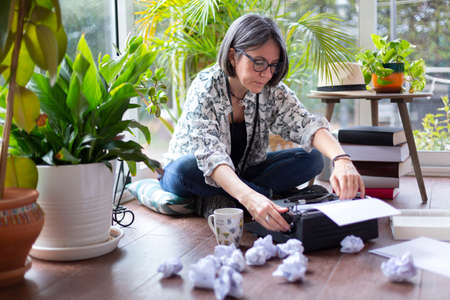 Adult female writer putting new blank sheet of paper on typewriter next to many crumpled papers. Creative block.の写真素材