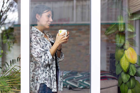 Adult woman with a cup of coffee in her hands looking thoughtfully through the window of her house. Time to think.の写真素材