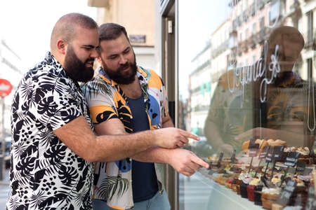 Young gay couple looking through the window of a pastry shop. Concept of everyday life in the city.の写真素材