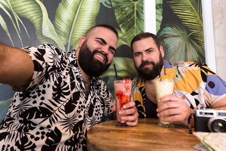 Happy young gay couple in summer clothes taking a self portrait together while having a cocktail on...