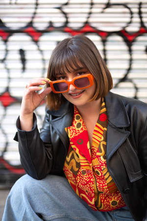 Young caucasian woman posing with style next to a graffiti wall. She wears a cute trendy outfit and sunglasses. Female outdoors portrait.の写真素材