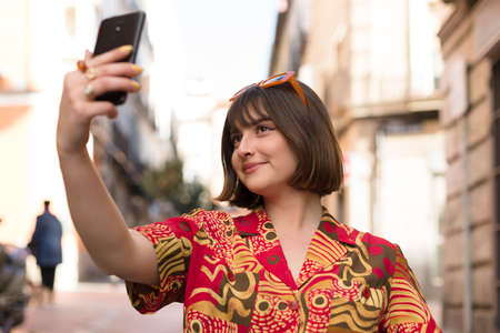Smiling young caucasian woman with short hair taking a photo of herself with a mobile phone outdoors.の写真素材
