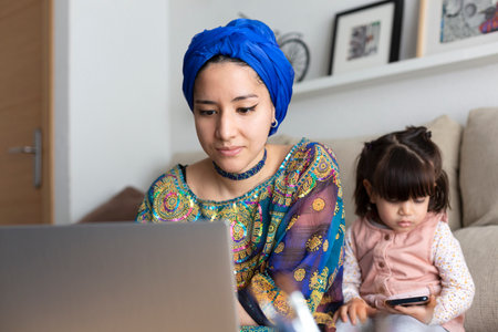 Closeup of young Muslim woman using laptop while her young daughter is watching something on mobile phone. Single parent family at home.の写真素材
