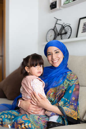 Young Muslim mother sitting on sofa with her little daughter in her arms. They are looking at the camera. Single parent family life.の写真素材