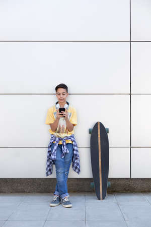 Caucasian teenage boy with skateboard using mobile phone. He is isolated on a wall. Space for text.の写真素材