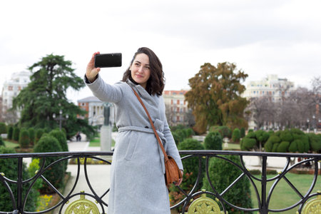 Young elegant woman sightseeing. She is taking a picture with her mobile phone of herself in a city garden. Space for text.の写真素材