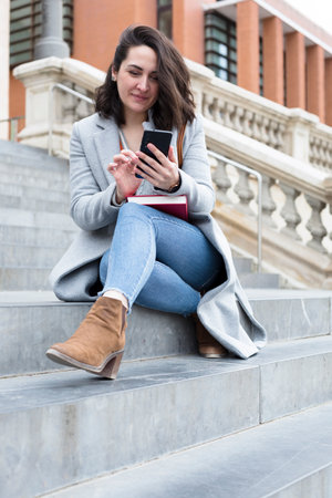 Elegant young caucasian woman using mobile phone. She is sitting on some steps outdoors.の写真素材