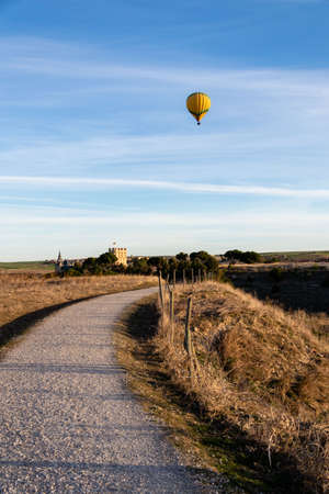 A road early in the day that heads towards a castle in the background. In the blue sky with tendrils of clouds a hot yellow hot air balloon stands out. Vertical photography.の写真素材