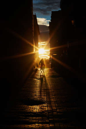 The silhouette of a cyclist outlined against the light advances down a cobbled street illuminated by the setting sun. Vertical photography. Copy space.の写真素材