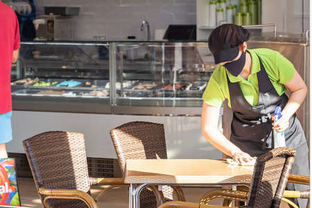 Huelva, Spain - July 4, 2020:  A waitress in face mask is cleaning a table with disinfectant liquid in the commercial center Islantilla. Protection against the coronavirus covid-19 in the new normalのeditorial素材