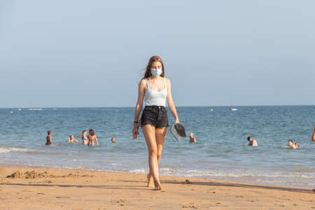 Punta Umbria, Huelva, Spain - August 7, 2020: A young Woman listening music is walking by the beach wearing protective or medical face masks. New normal in Spain with social distancingのeditorial素材