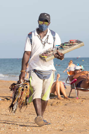 Punta Umbria, Huelva, Spain - August 7, 2020: African immigrant selling their wares on the beach wearing protective or medical face mask. All goods of these vendors are brand copiesのeditorial素材