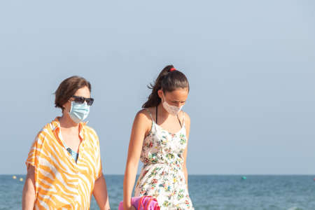 Punta Umbria, Huelva, Spain - August 7, 2020: Mother and daughter walking by the beach wearing protective or medical face masks. New normal in Spain with social distancingのeditorial素材