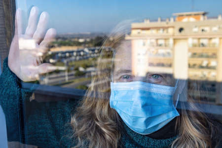Woman isolated by coronavirus Covid-19 is looking through the glass of her house wearing a protective face maskの写真素材